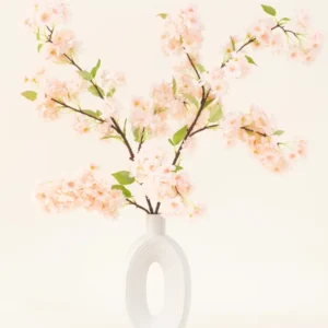 Eine weiße Keramikvase mit einer breiten ovalen Öffnung zeigt mehrere grünblättrige künstliche Seidenblumenstrauß hellrosa Kirschblütenzweige vor einem einfachen hellen Hintergrund.